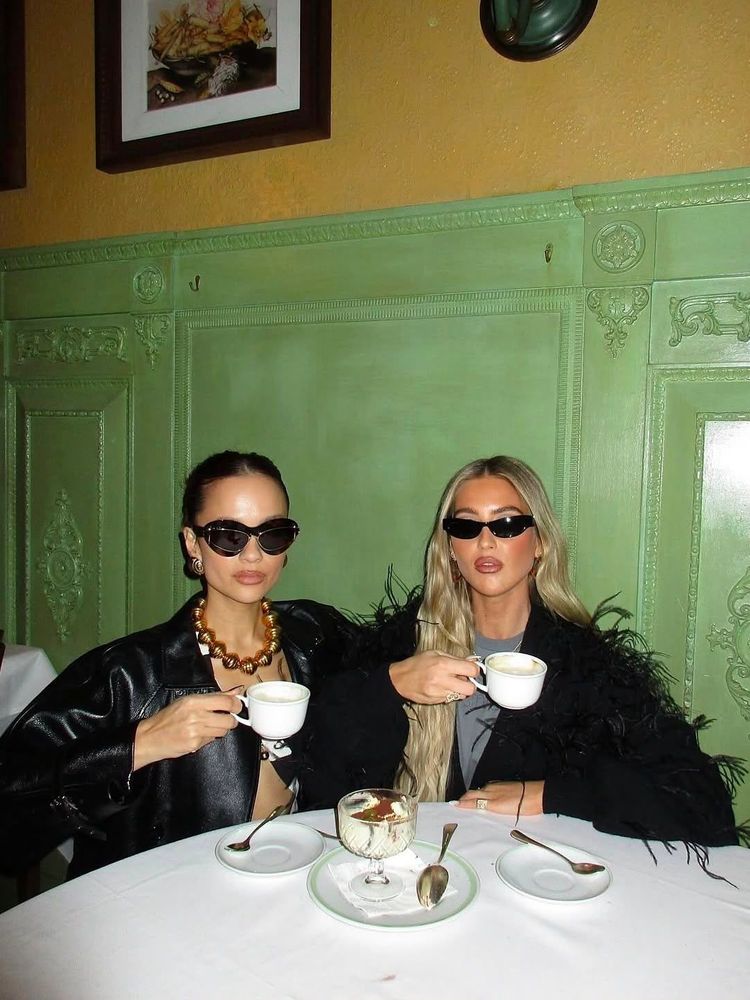 Emili Sindlev with friend drinking coffee wearing both black sunglasses and black jackets, the café has yellow and green walls with cornice, the table has a white cloth and dishes