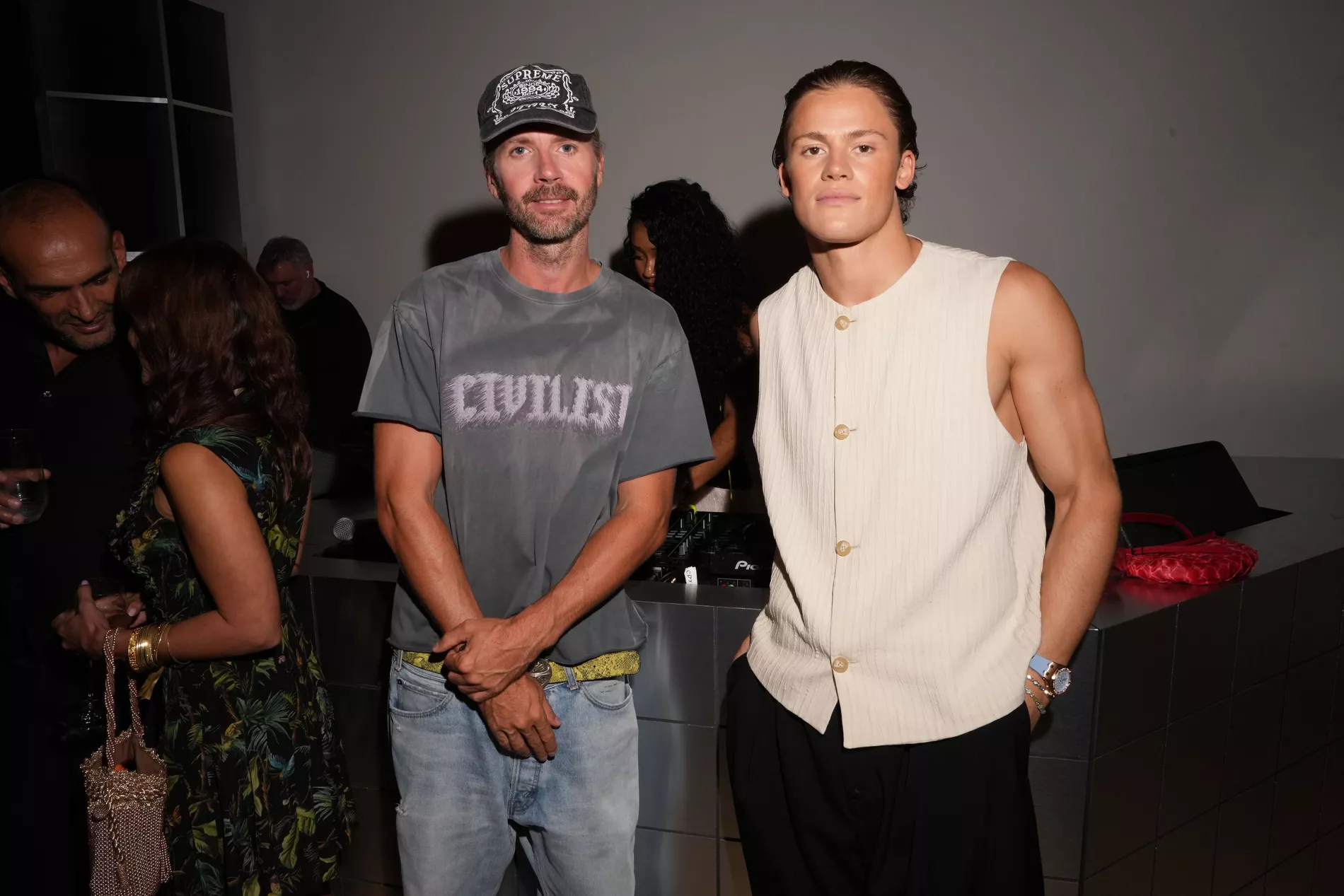 Two men standing indoors near a counter, with the man on the left wearing a dark gray Civilist t-shirt, light-wash jeans, and a cap, and the man on the right wearing Emporio Armani Embossed Jacquard Fabric Waistcoat with Wrinkle Effect.