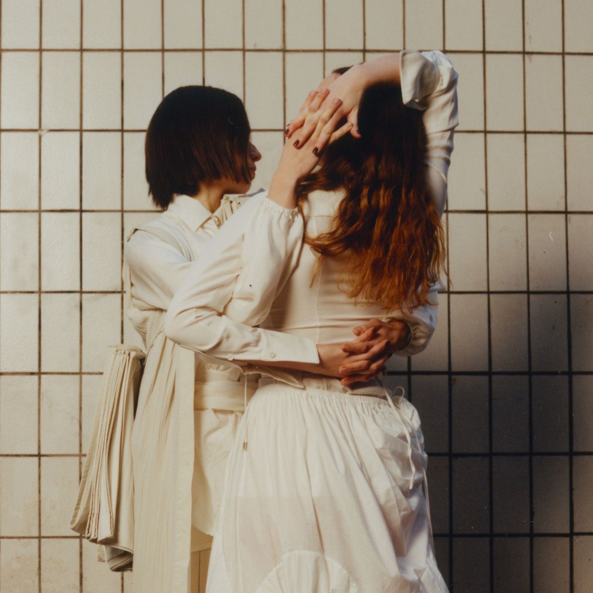 Two women holding hands behind their backs in a tiled room wearing all white