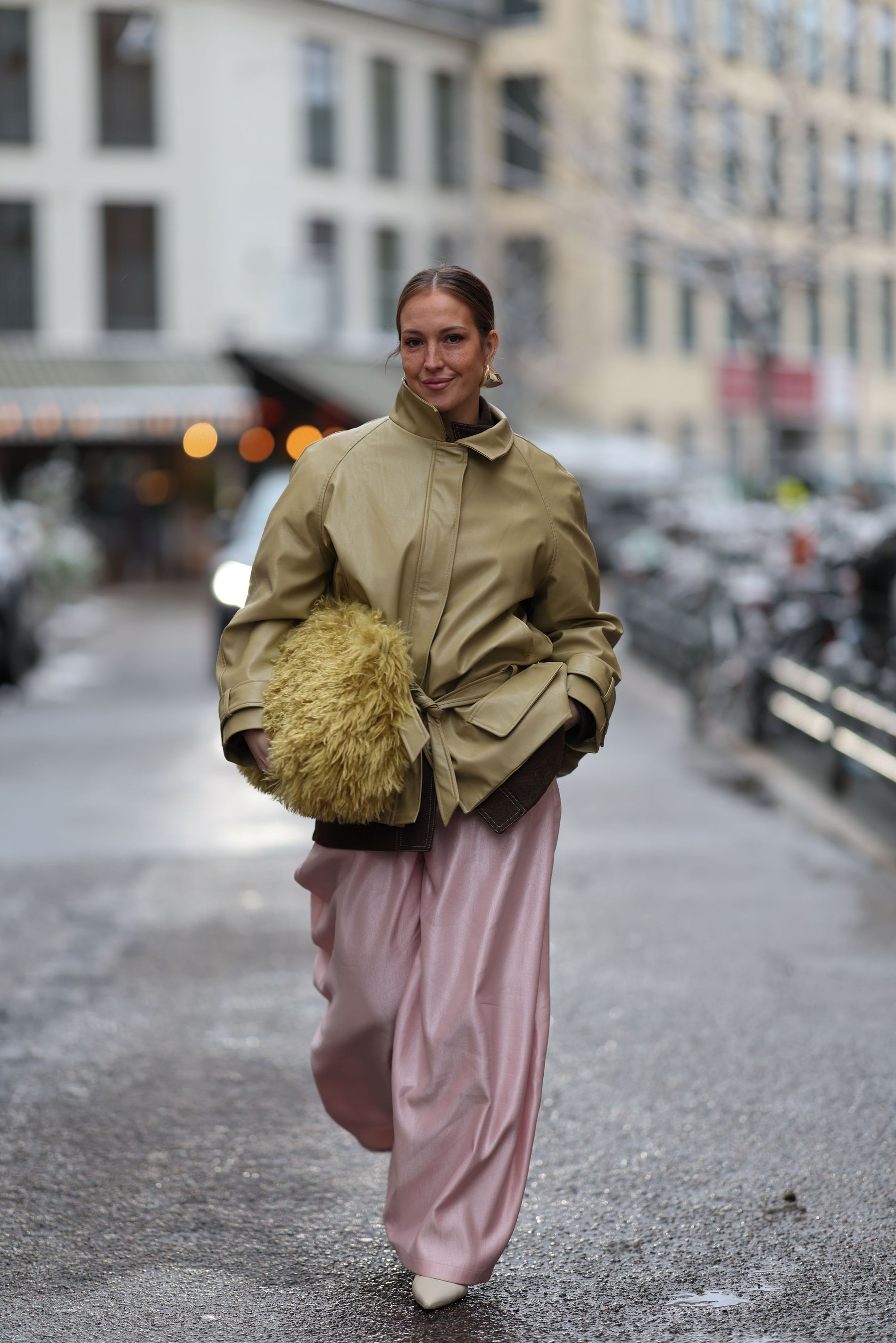 Streetstyle images of a women walking with a beige belted leather jacket with, paired with loose pink trousers and a green big fluffy bag