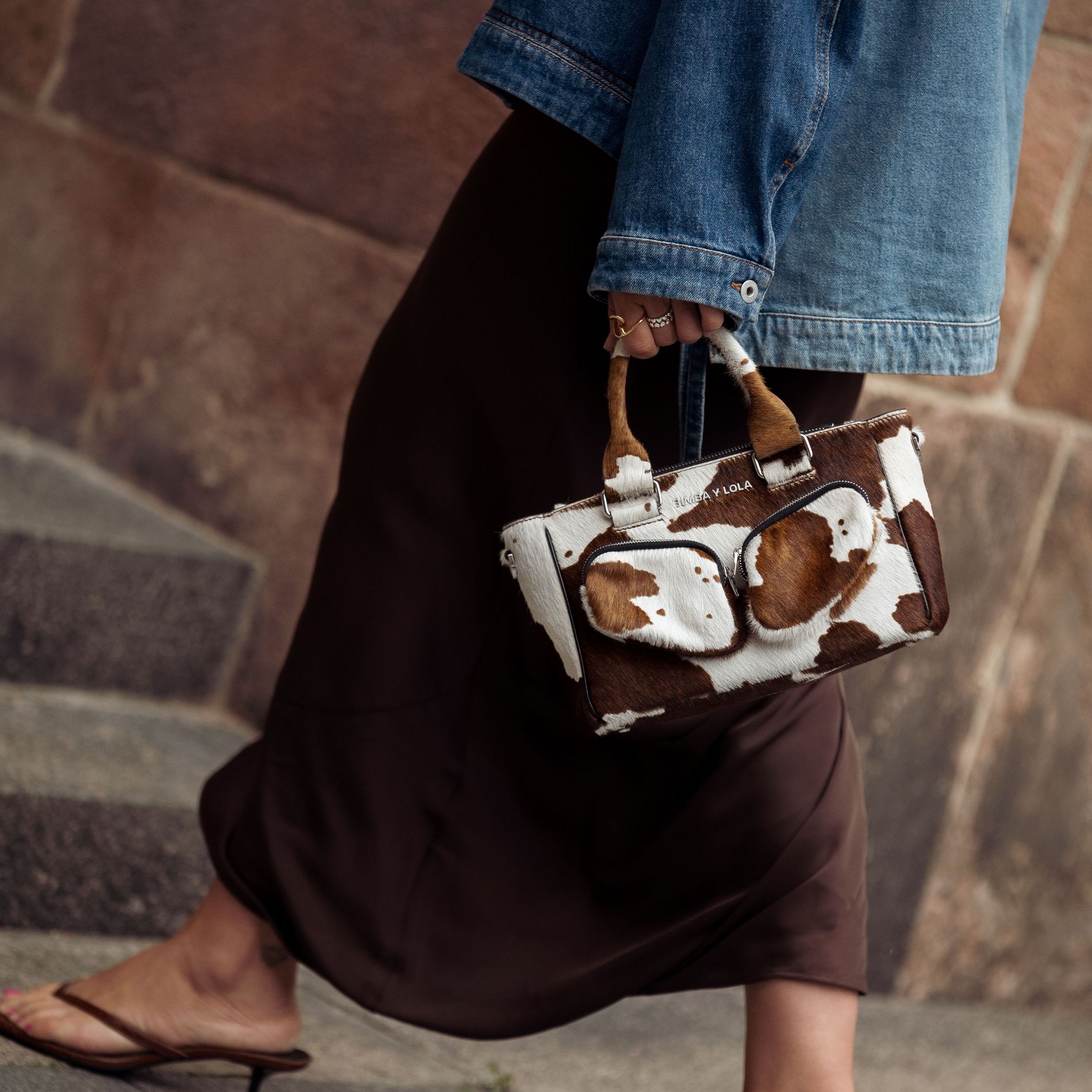 Close-up of a brown and white cow print mini bag styled with a brown slip skirt, kitten heels, and a denim jacket
