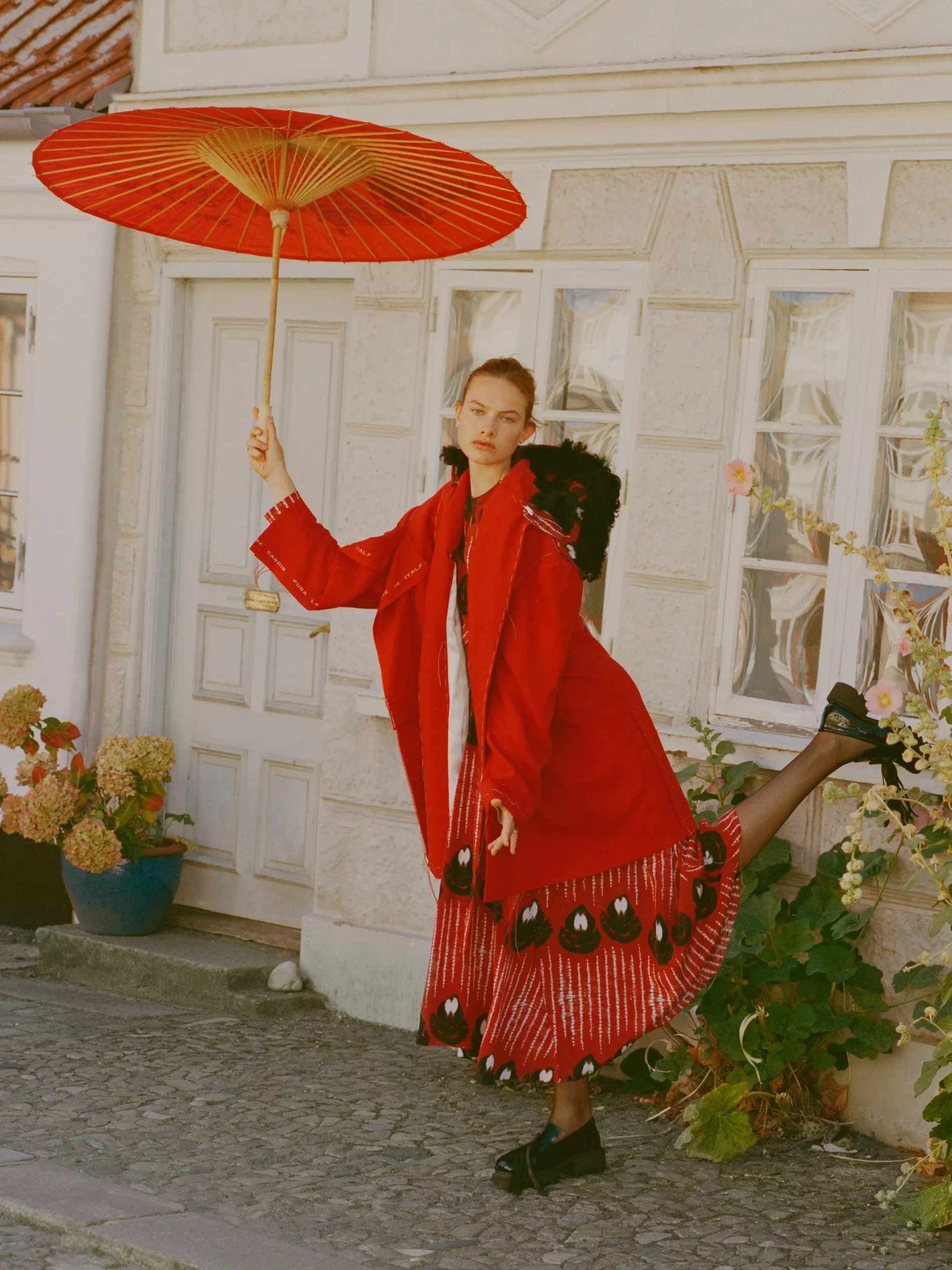Model stands in a courtyard in a red coat, red skirt and black shoes holding a red umbrella