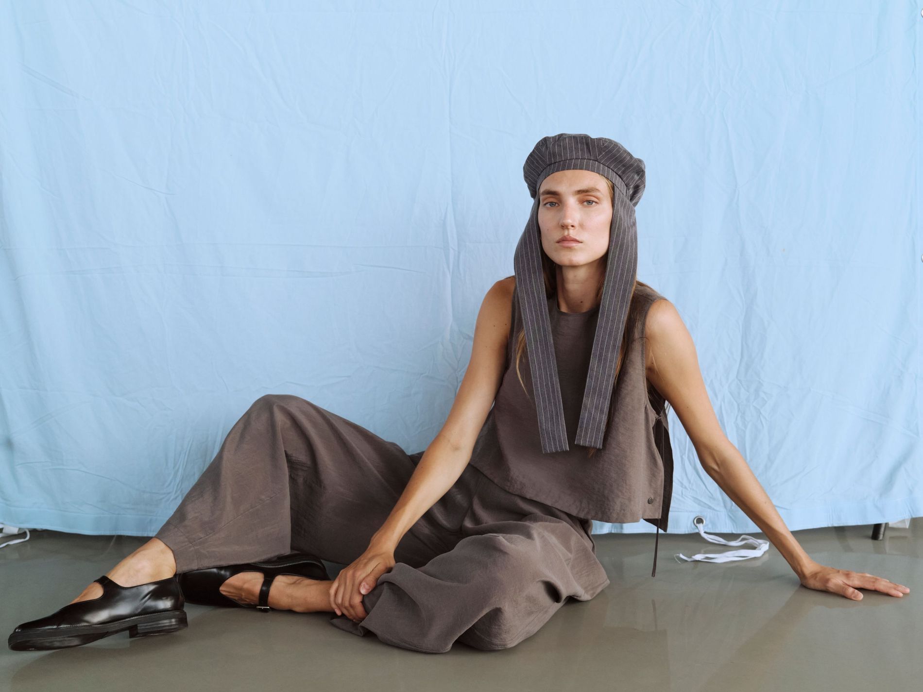 Model poses in a pinstriped hat, greige cotton top, trousers and black leather Mary janes 