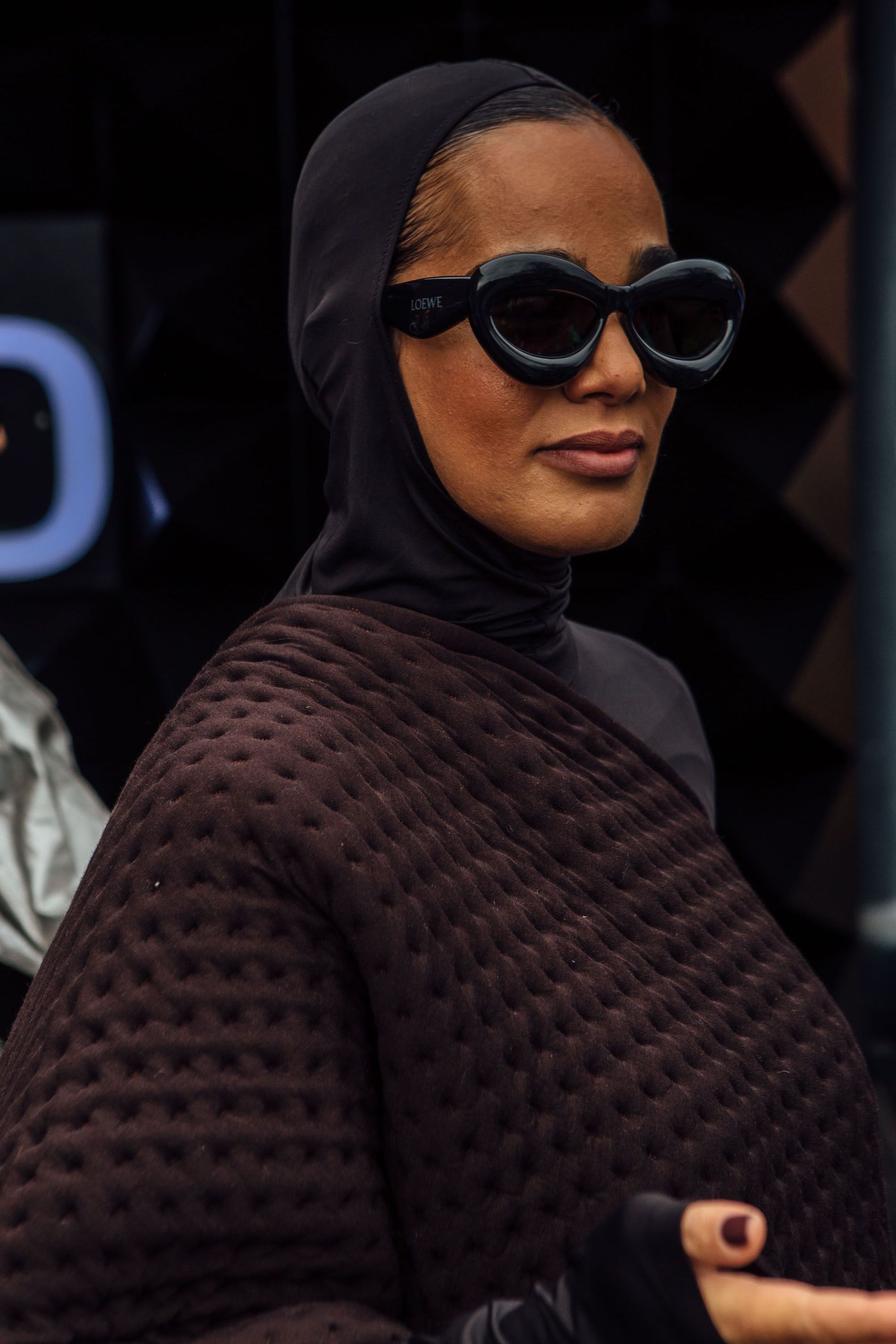 A guest wears black Loewe bubble sunglasses and a brown wrap