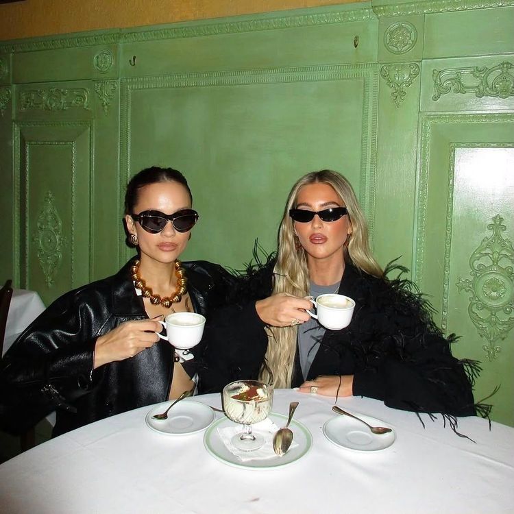 Emili Sindlev with friend drinking coffee wearing both black sunglasses and black jackets, the café has yellow and green walls with cornice, the table has a white cloth and dishes