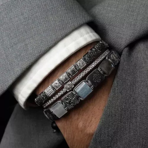 Three luxury braided and beaded Shamballa Jewels bracelets featuring dark metallic elements, square bead stations, and diamond or gemstone accents worn against a grey jacket cuff mans wrist, the mana is wearing a grey suit and a white shirt 