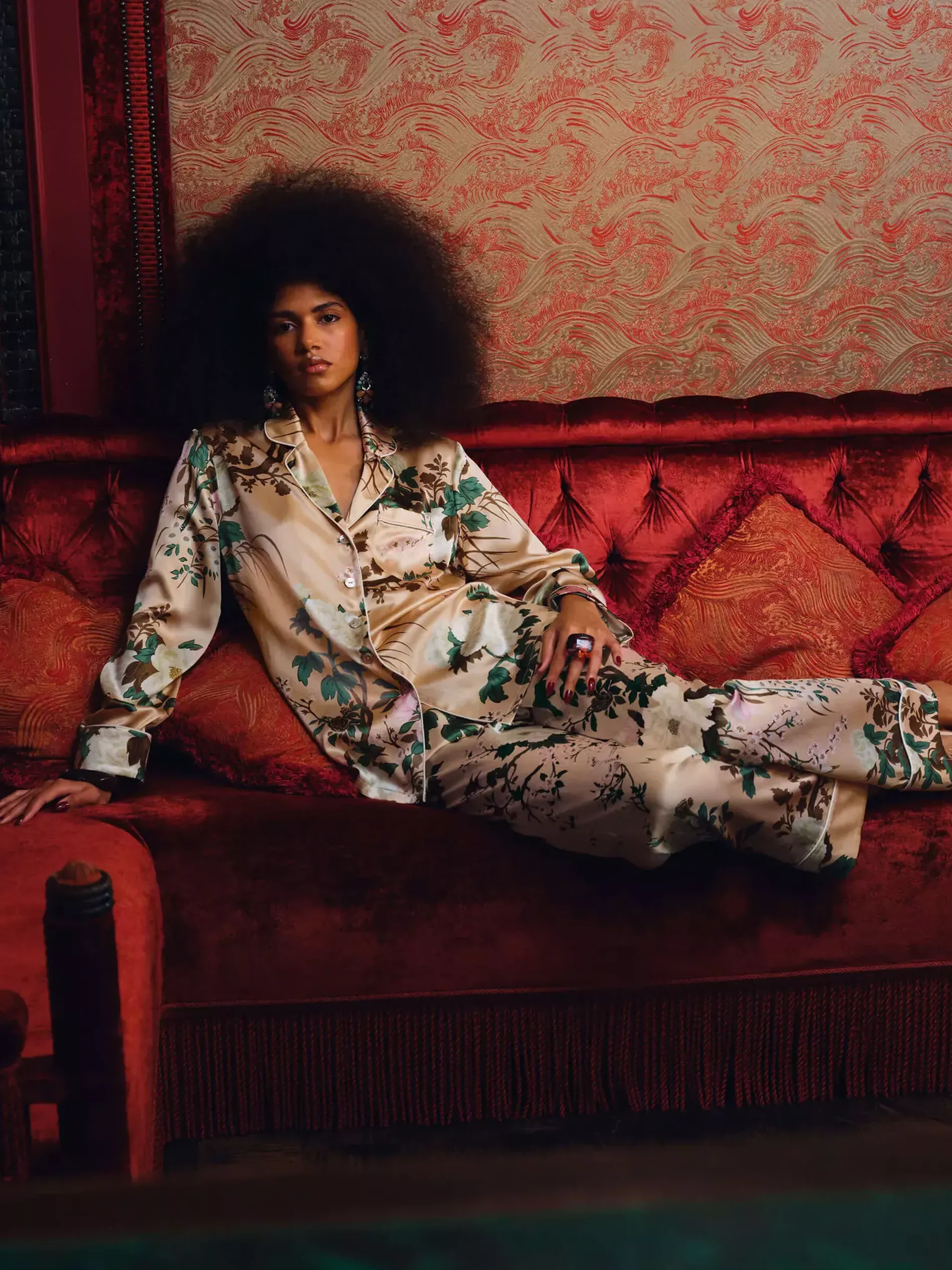 Woman leaning on a red velvet sofa wearing a patterned silk pyjama in pink with light pink feather shoes 