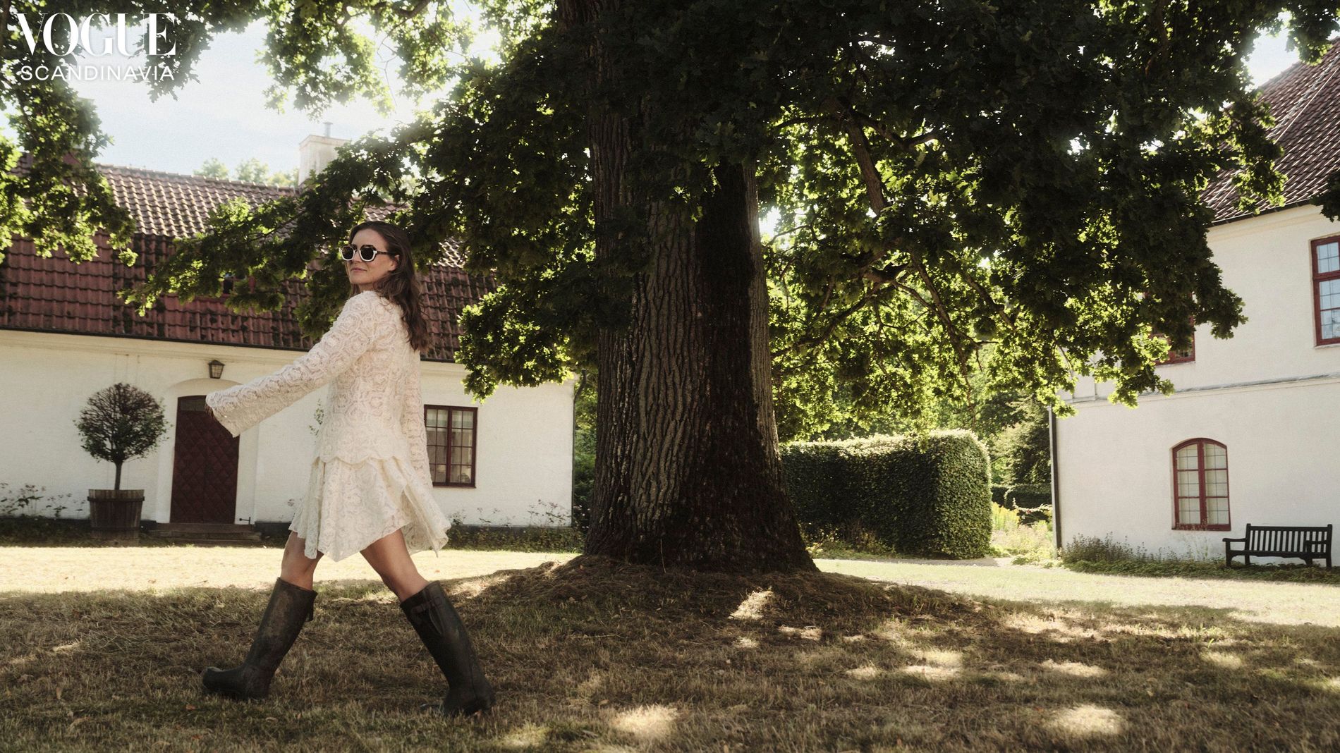Beate Heuman walks across a yard with two white buildings with tile roofs in the background and a tree in centre frame. She wears a lace mini dress with long sleeves, sunglasses and wellington boots