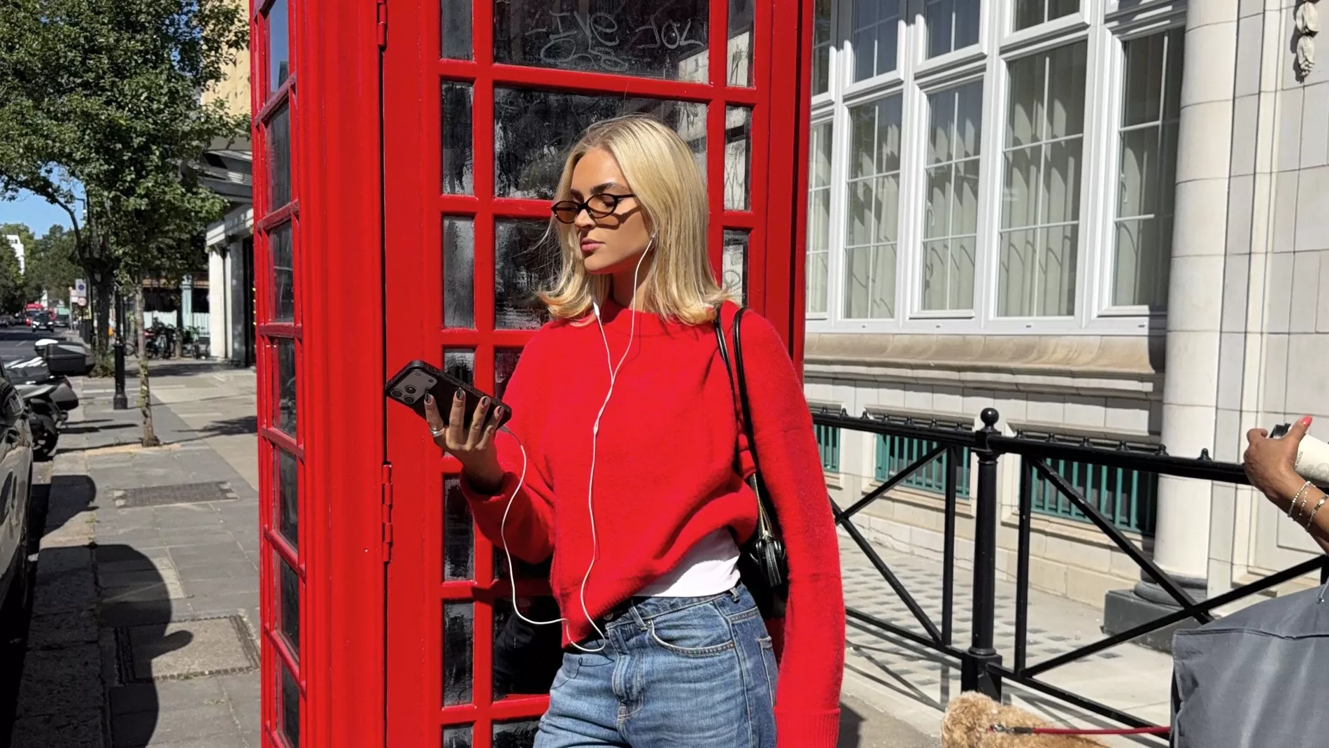 Emilia wears balck flipflops, jeans, and a red jumper by a London telephone box 