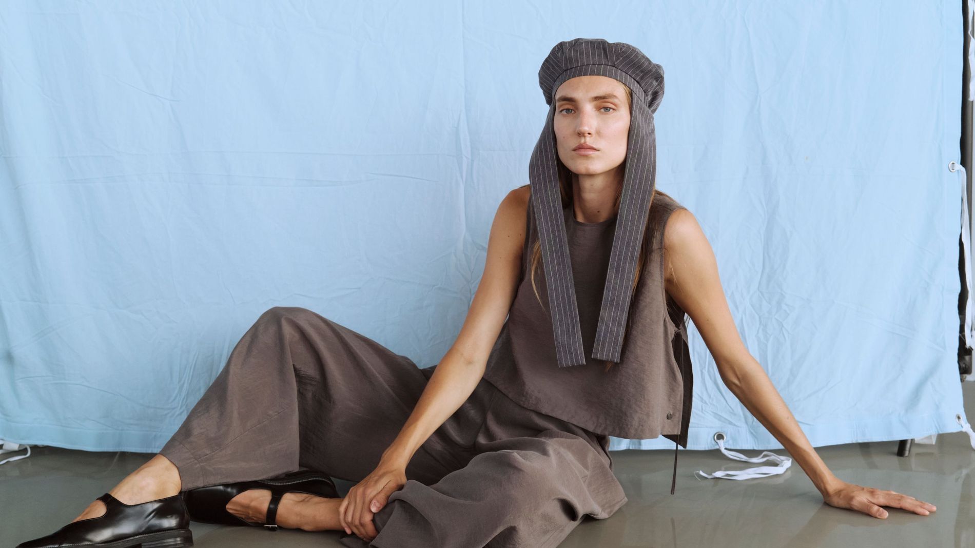 Model poses in a pinstriped hat, greige cotton top, trousers and black leather Mary janes 