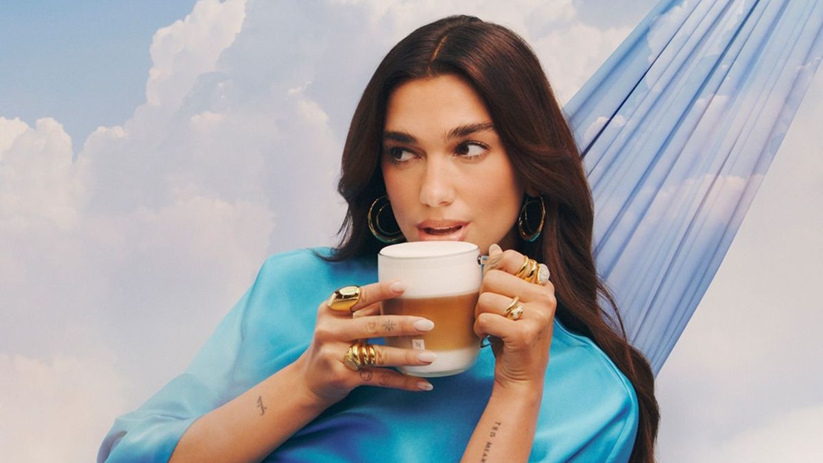Dua Lipa announced as Nespresso's brand ambassador. The image sees her suspended in the clouds, in a hammock, a cup of frothy coffee in her hand. She is dressed in a blue satin gown from Pinko