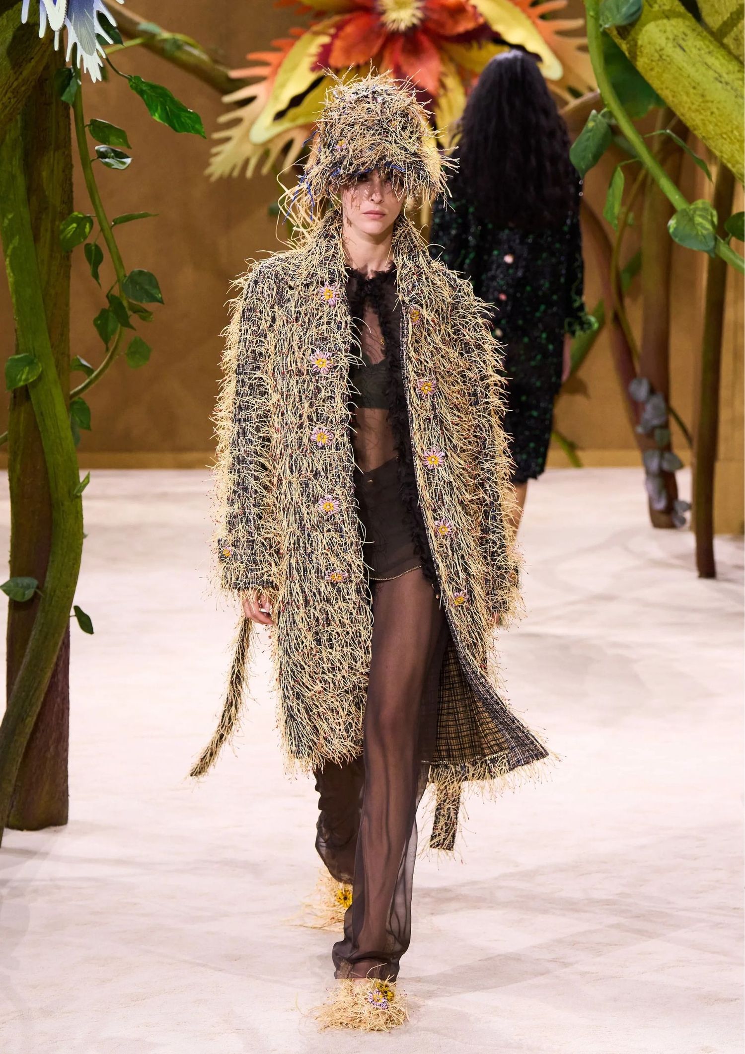 Long textured coat with a matching oversized fringed hat, a sheer long-sleeved top layered over a black bralette, sheer black tights, and textured shoes.