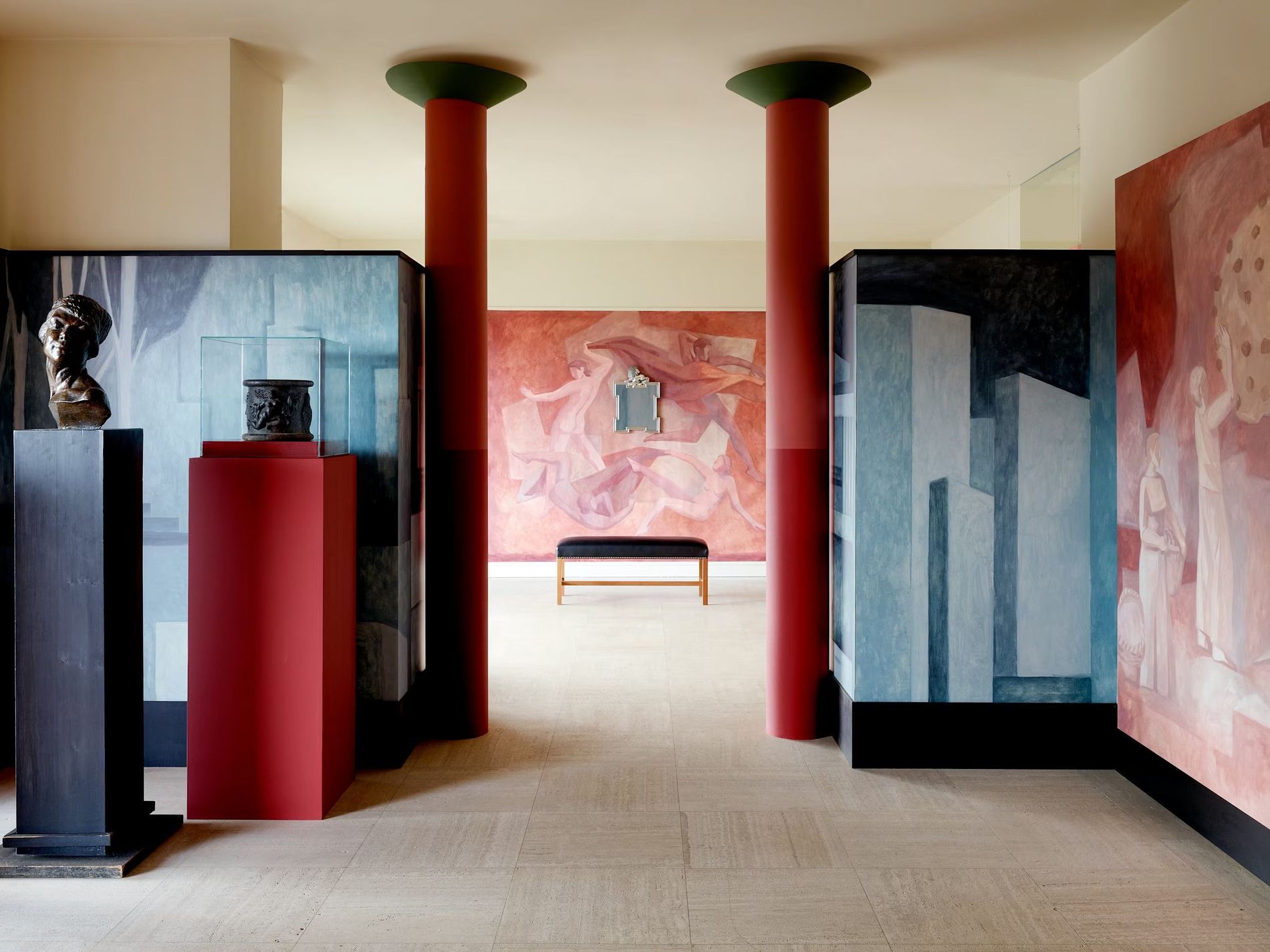A gallery-like interior from Svenskt tenn Fall' 26featuring two tall red columns with dark green tops, surroundeed by black and blue display panels, sculptural objects, abstract paintings in red, pink and blue tones and a smal bench in the middle