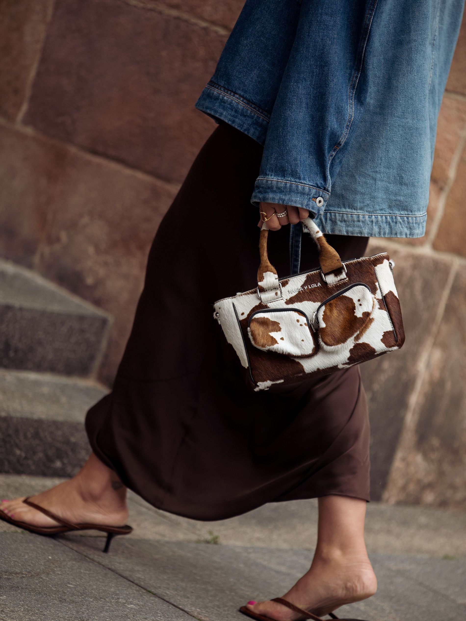 Close-up of a brown and white cow print mini bag styled with a brown slip skirt, kitten heels, and a denim jacket