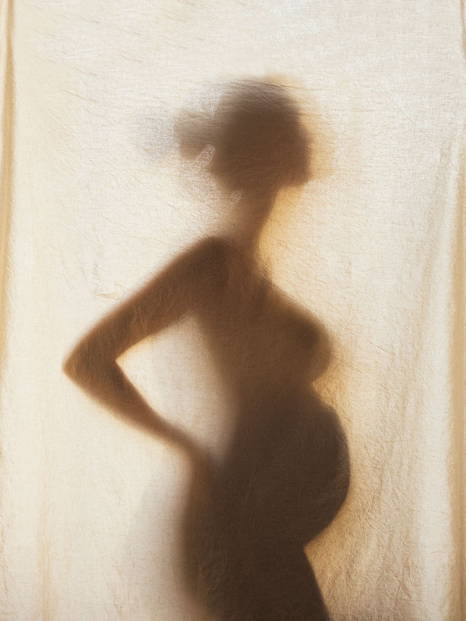 Silhouette of a pregnant woman is seen through a linen curtain