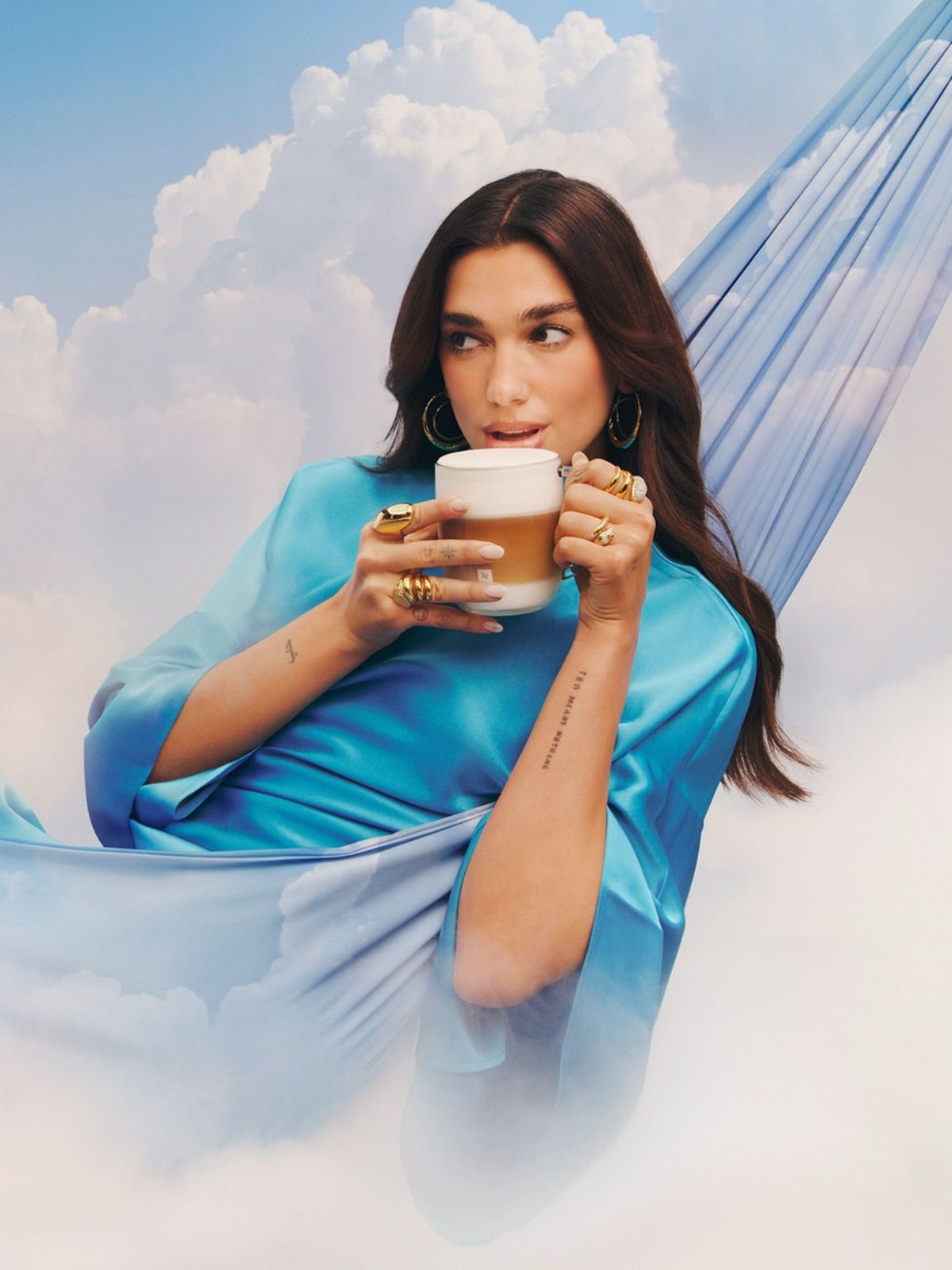 Dua Lipa announced as Nespresso's brand ambassador. The image sees her suspended in the clouds, in a hammock, a cup of frothy coffee in her hand. She is dressed in a blue satin gown from Pinko