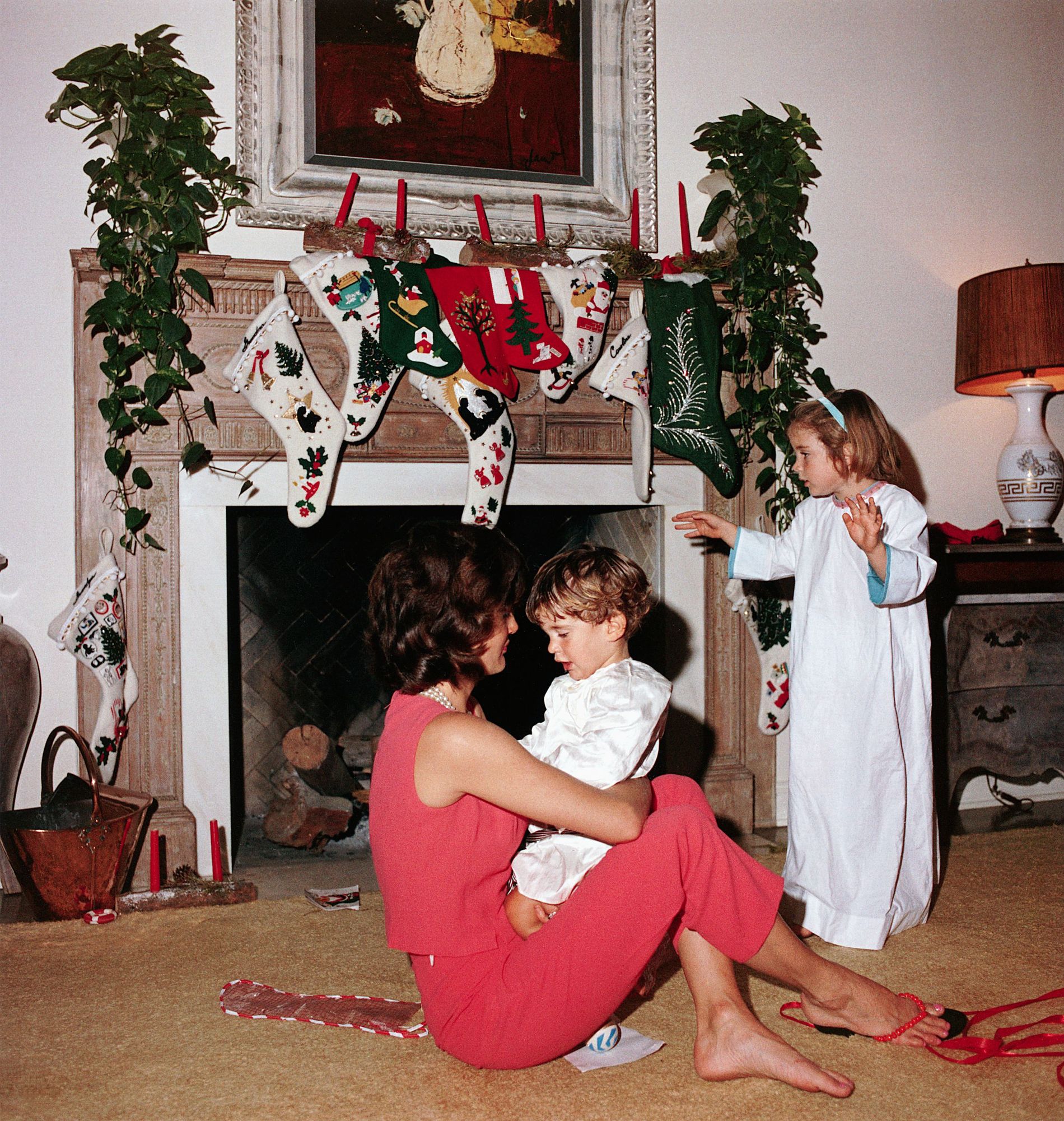 Jacqueline Kennedy with the children on Christmas morning. She sits on the floor, holding John on her lap, and Caroline stands nearby in her nightgown. Several stockings hang from the mantle.