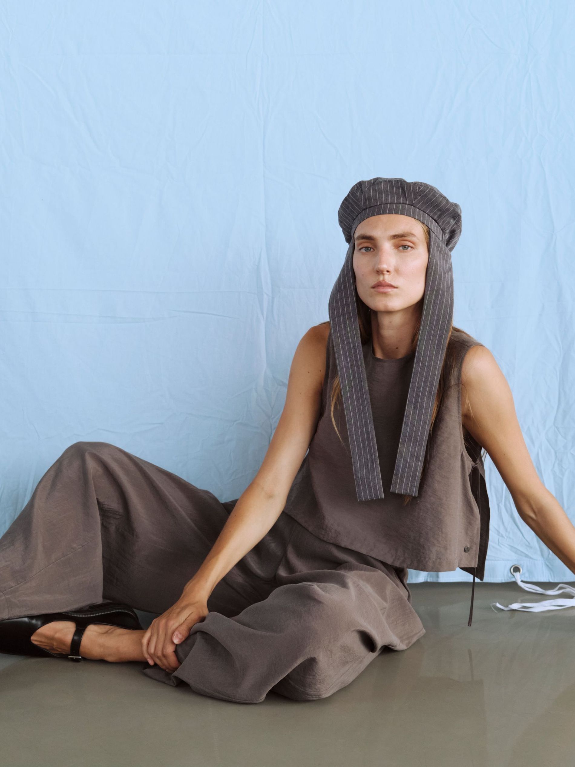Model poses in a pinstriped hat, greige cotton top, trousers and black leather Mary janes 