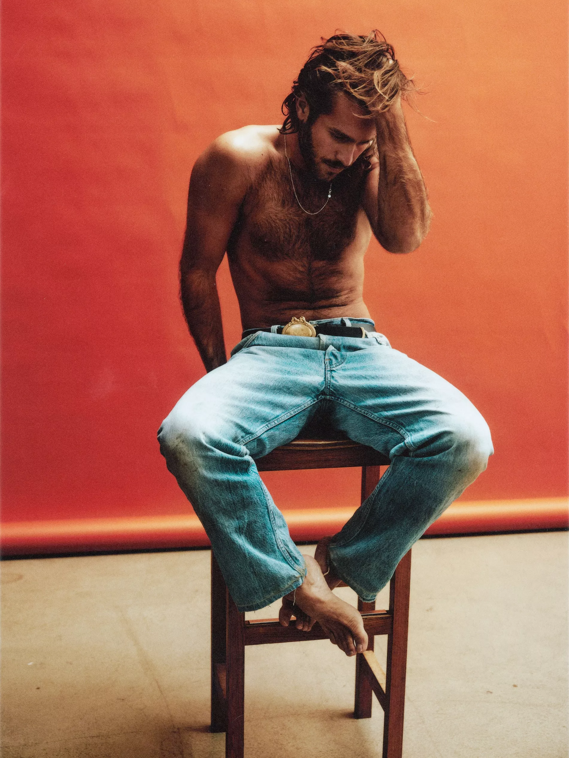 Benjamin Ingrosso sitting on a high stool, shirtless and wearing jeans, the background is bright orange