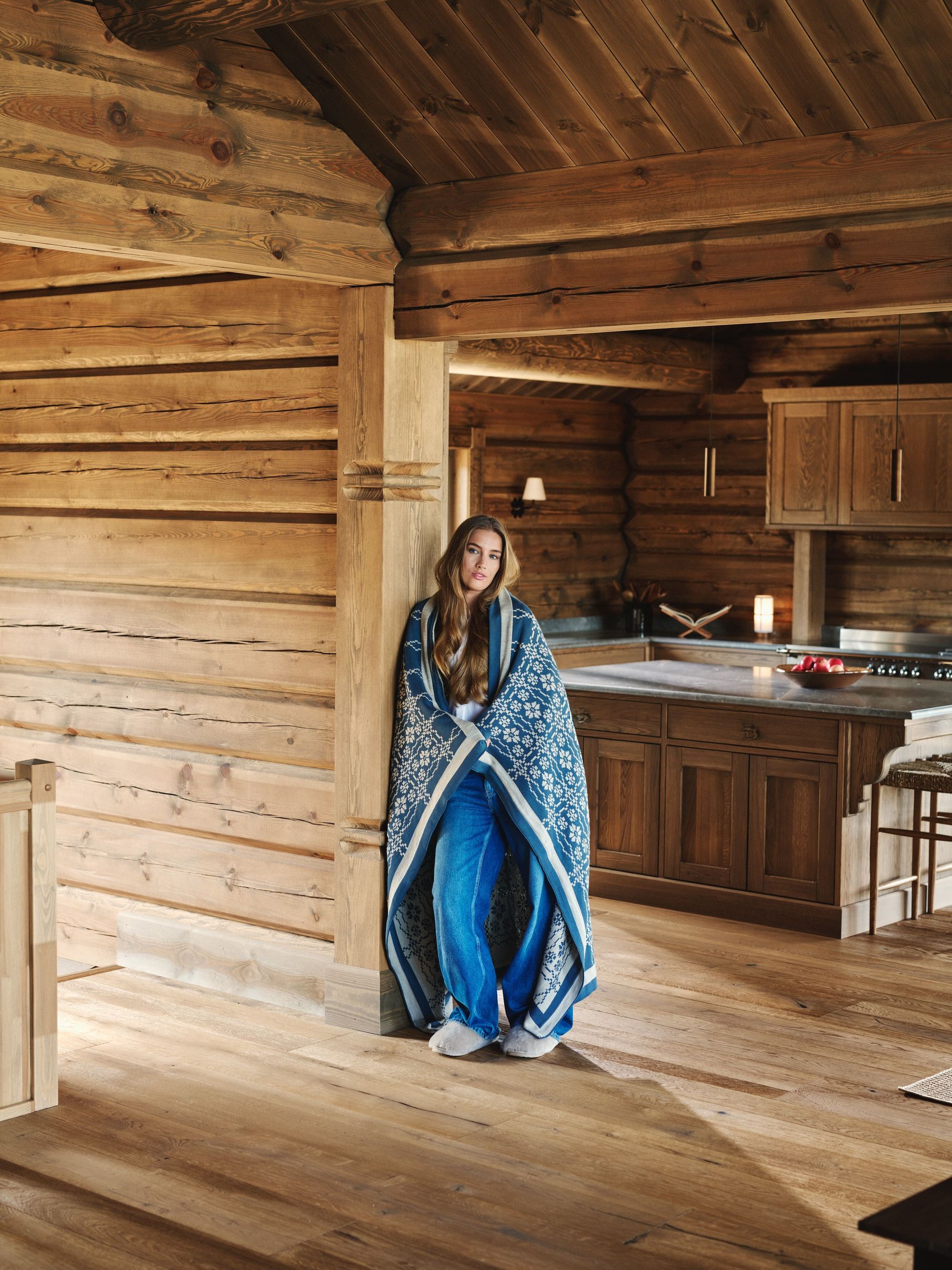Pernille Døsvik leaning against a wall in a wooden house wearing on of her blankets from the collaboration with Dale Of Norway