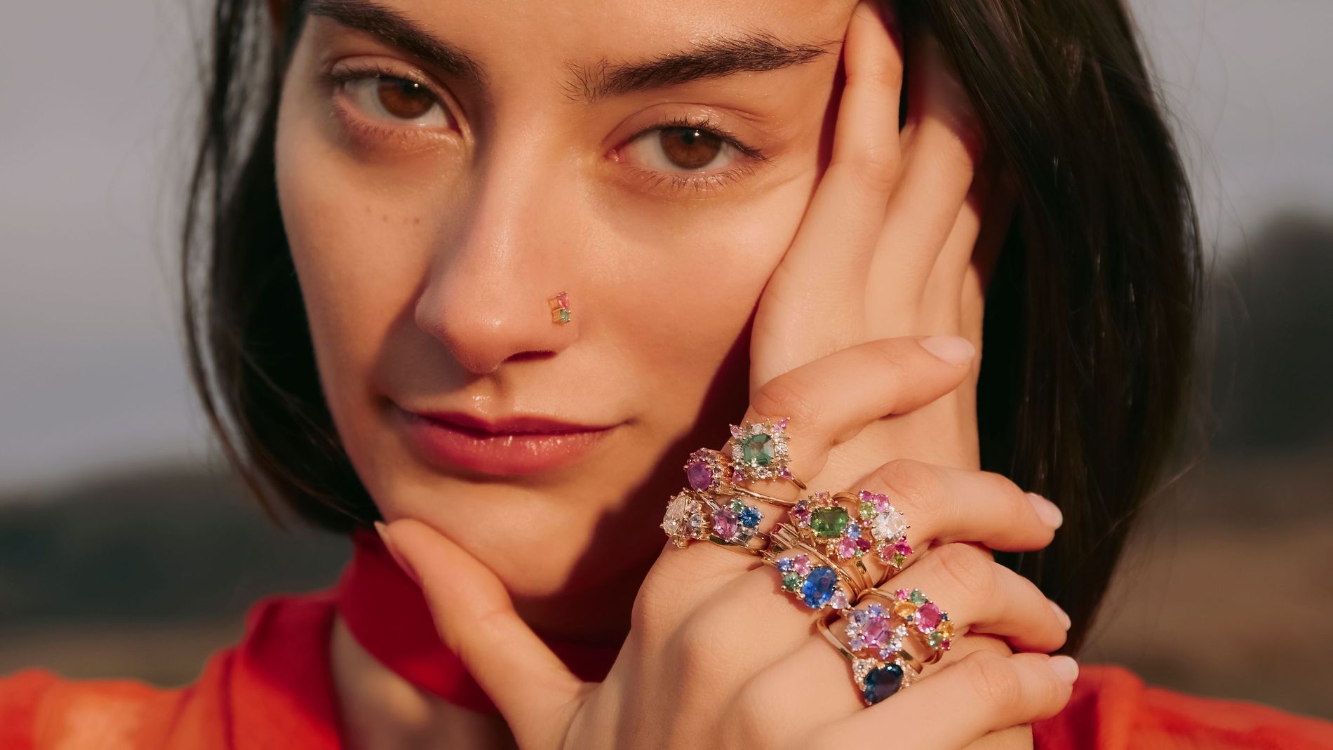 Up-close model posing with resting hands decorated with colourful gemstones from Anpé Atelier she is wearing a red-orange loose fitting top