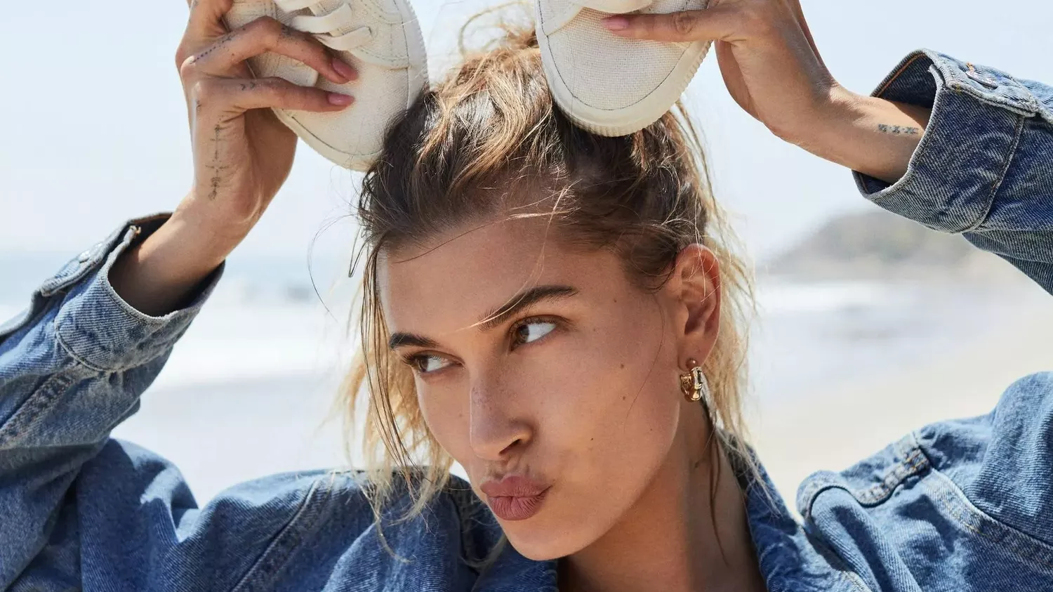 Hailey Bieber standing on a beach wearing a jean jacket and white top with white sneakers on top of her head