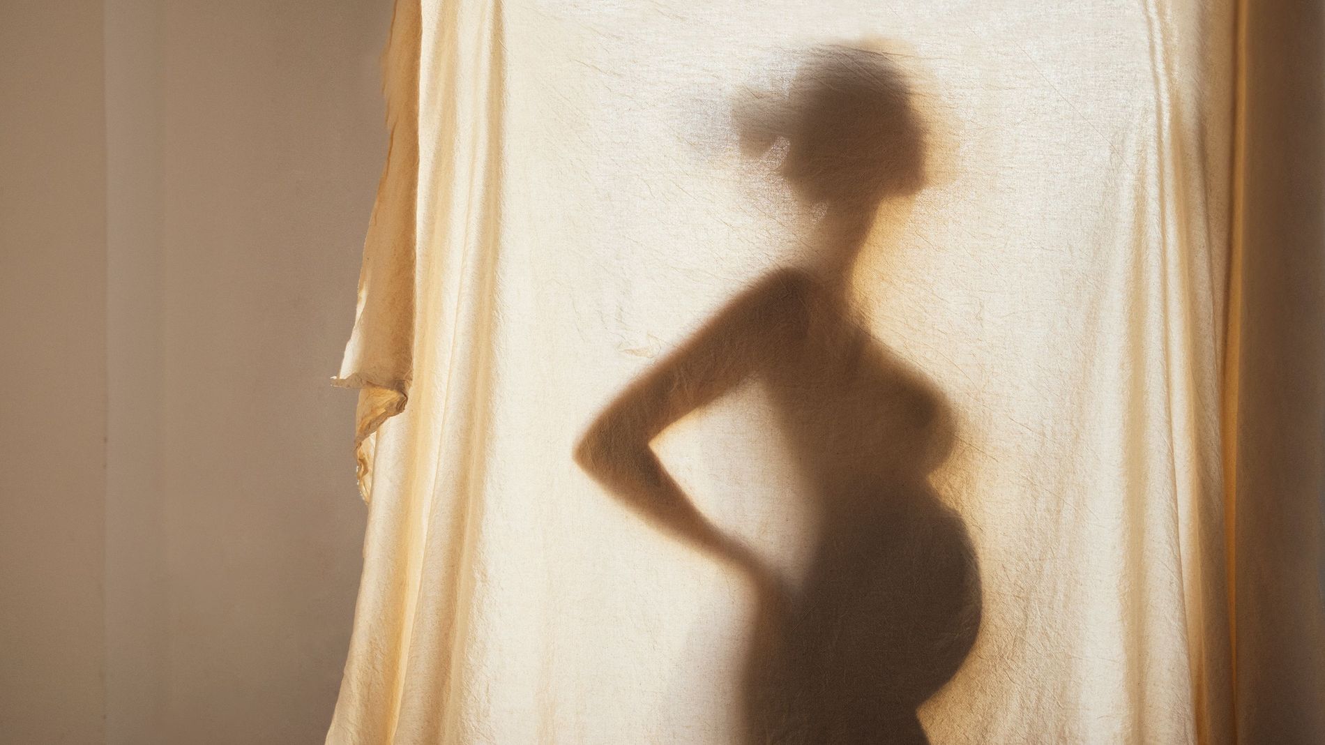 Silhouette of a pregnant woman is seen through a linen curtain