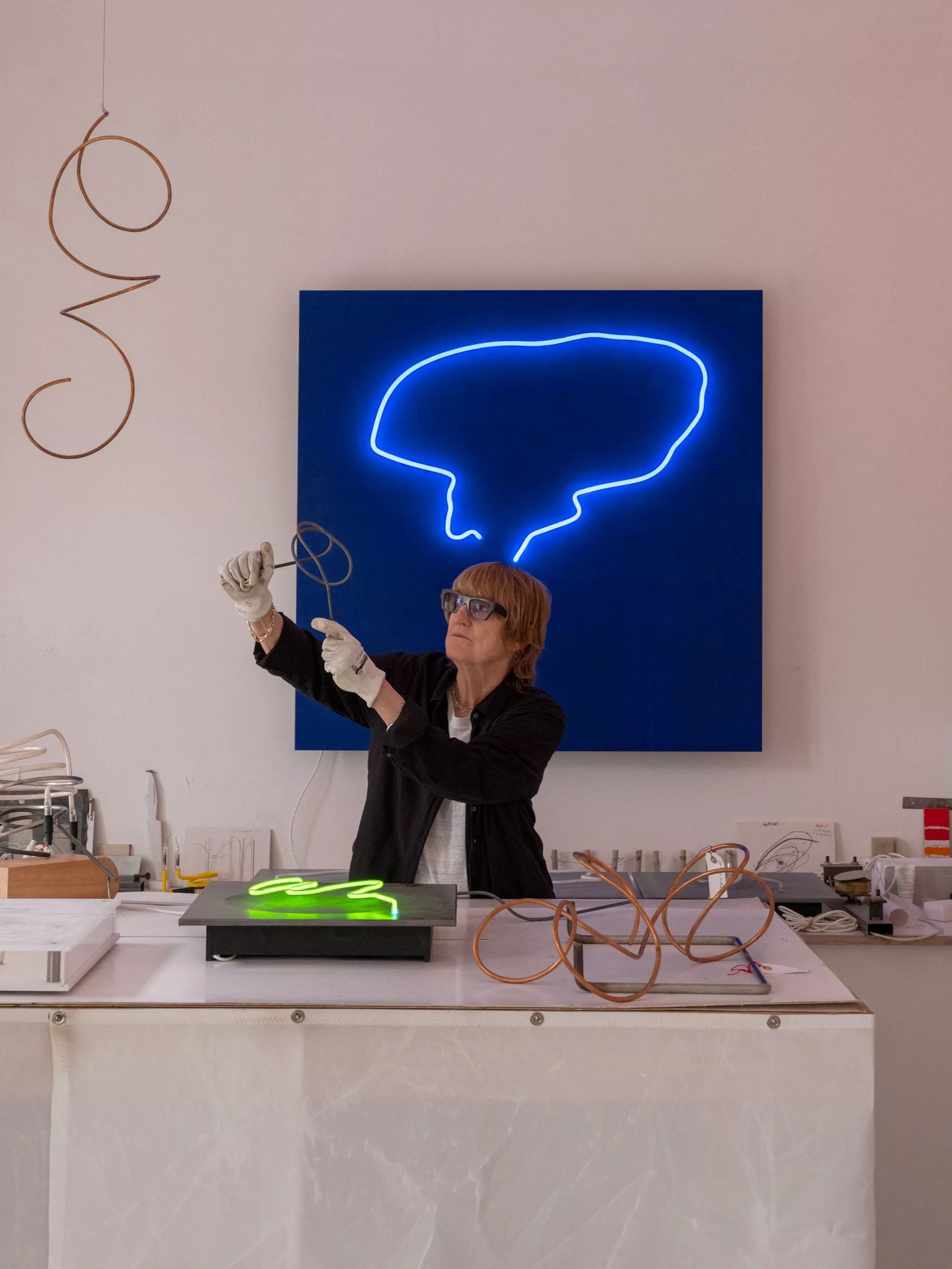 Gun Gordillo standing behind a desk with exam glasses working on a new creation, behind her is a dark blue painting and a neon blue loop shines