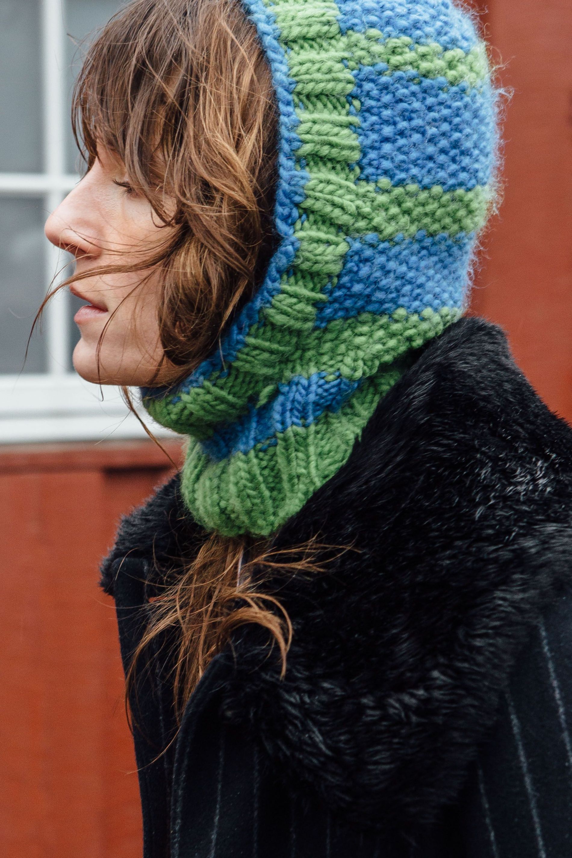 A street style CPHFW AW23 guest wears a green and blue patterned balaclava, and pin-striped coat with a faux fur collar