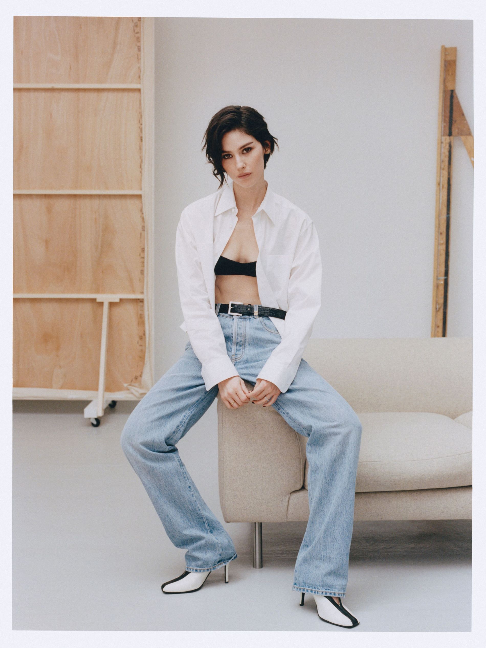 Gracie Abrams poses in a white shirt, black bralette, belted light wash jeans and black and white pumps on a soundstage with a beige couch