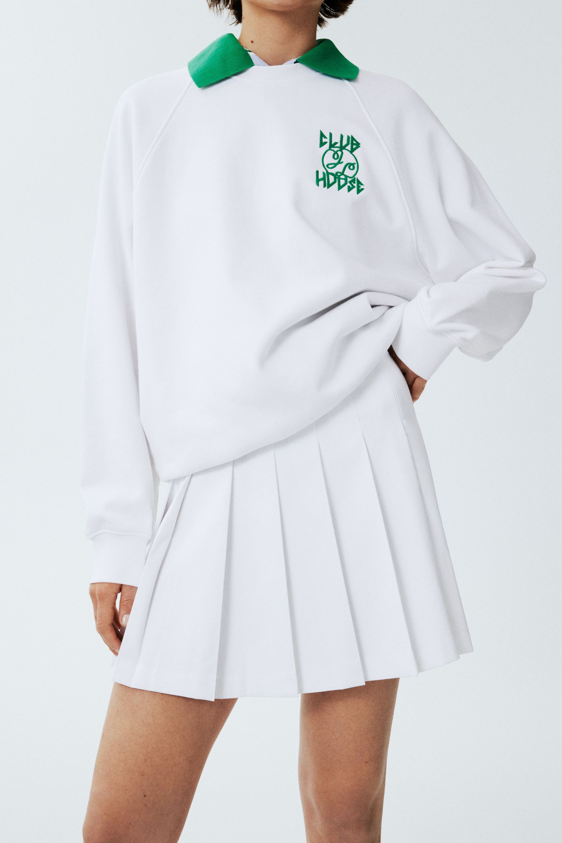 A white collared sweatshirt with green details and a white pleated tennis skirt from J.Lindeberg's Tennis 2024 collection